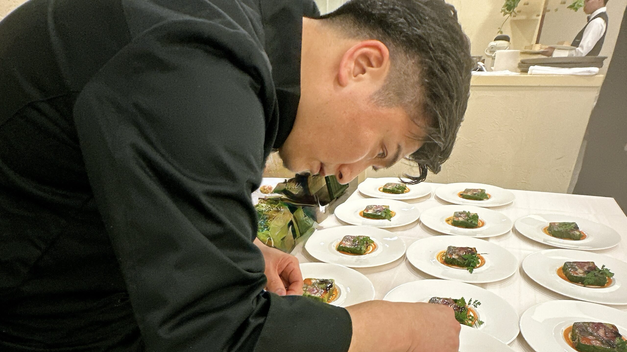 Un chef en veste noire termine des terrines avec des herbes sur des rangées d’assiettes blanches